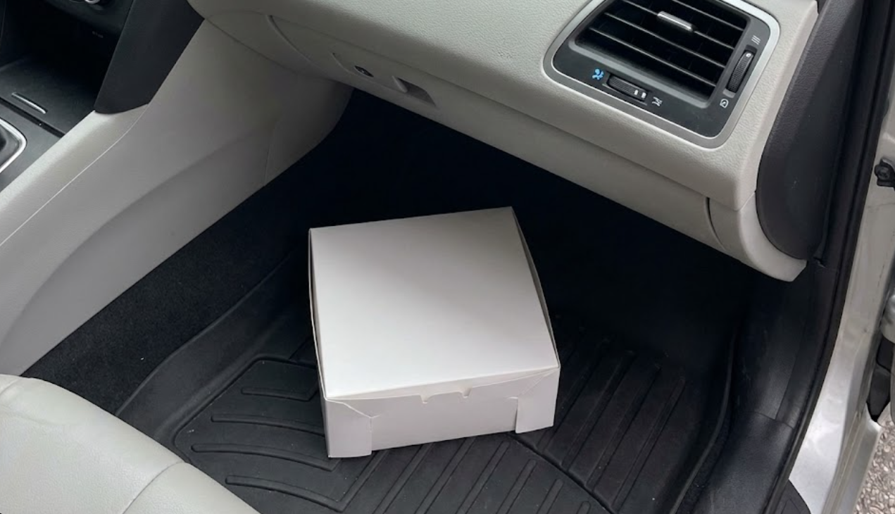 White cake box placed securely on the flat floor of a car's passenger seat footwell.
