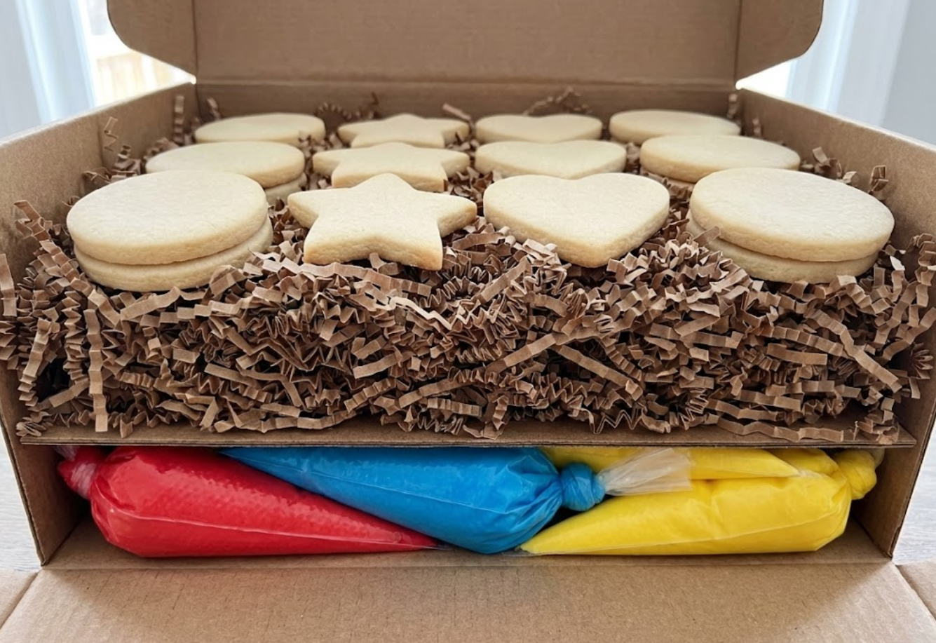 Cross-section cutaway view of a packed DIY cookie decorating kit box, showing the proper layering technique: heavy icing bags at the bottom foundation, a thick middle layer of crinkle paper cushion, and plain sugar cookies nestled on top.