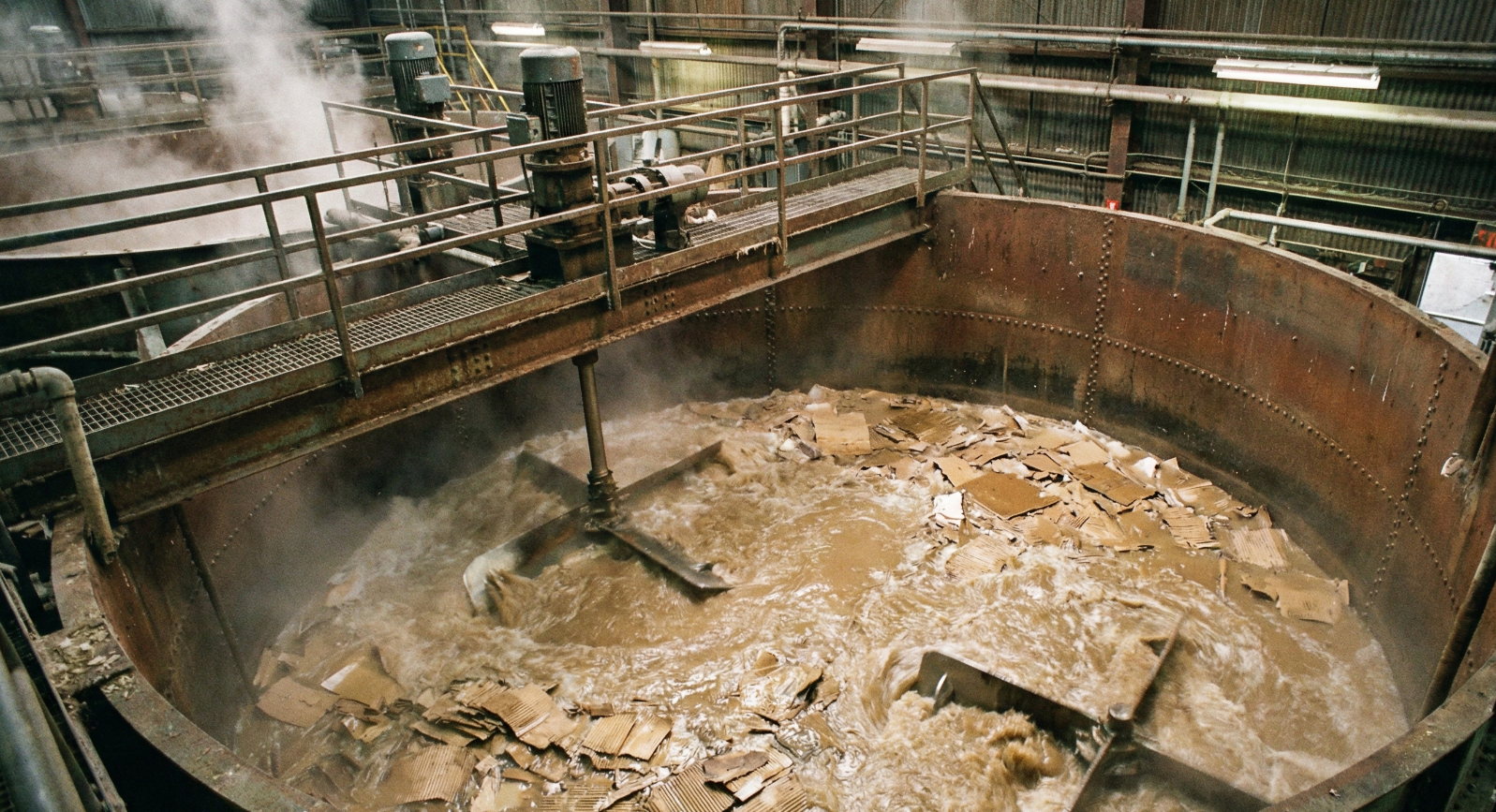 An overhead view of a large industrial hydropulper vat churning warm water and shredded cardboard boxes into a thick brown fiber slurry.
