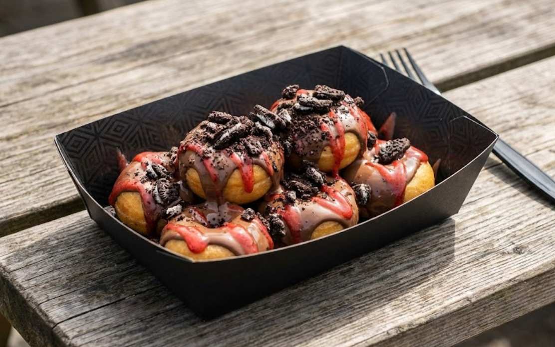 Matte black greaseproof paper food tray holding loaded mini donuts with chocolate and Oreo toppings, sitting on a wooden table with no visible grease stains.